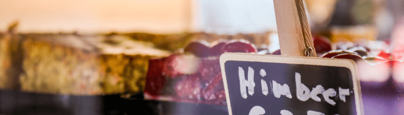 Ein Ausschnitt der Kuchentheke mit Obst und Rührkuchen. Vor einem Blech Himbeerkuchen steht ein handbeschriebenes Schild mit dem Wort "Himbeer"