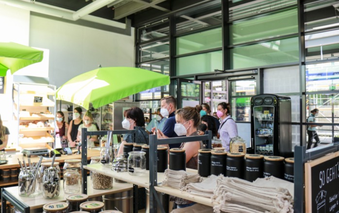 Der Laden mit Besuchern bei der Eröffnungsfeier. Alle tragen den vorgeschriebenen Mund-Nasen-Schutz. Man sieht die unterschiedlich gefüllten Regale und den gefüllten Kühlschrank.