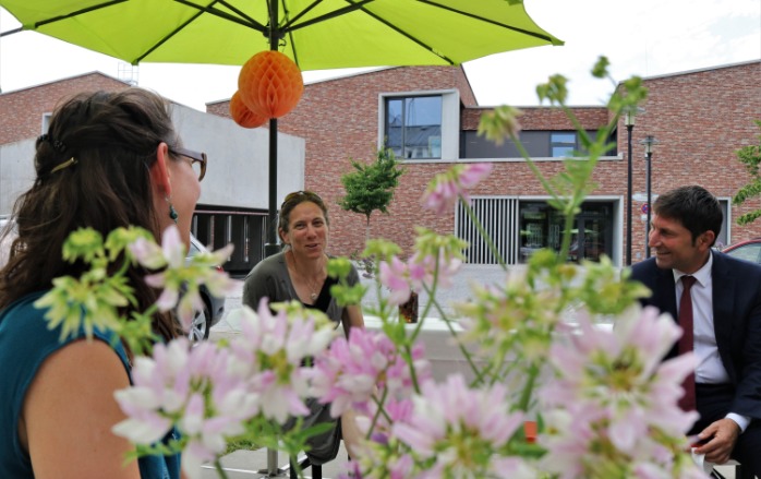 Drei Personen, darunter auch der Unterföhringer Bürgermeister Herr Kemmelmeyer unterhalten sich auf den festlich geschmückten Biertischen mit Wiesenblumen und orangen Papierdekokugeln, die an grünen Sonnenschirmen hängen, vor dem Laden bei der Eröffnungsfeier