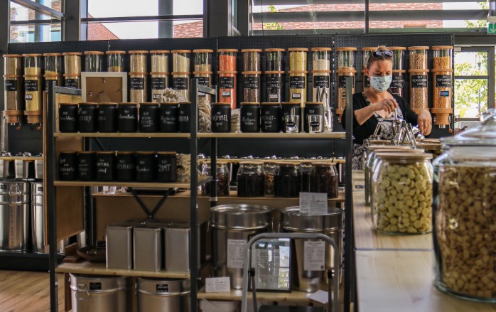 Gefüllte Regale, in der letzten Reihe befinden sich die großen Bins mit dem trockenen Schüttgut. Eine Besucherin füllt gerade ein Produkt in einen Behälter, sie trägt den vorgeschriebenen Mund-Nasen-Schutz. Es ist am Tag der Eröffnungsfeier aufgenommen worden.